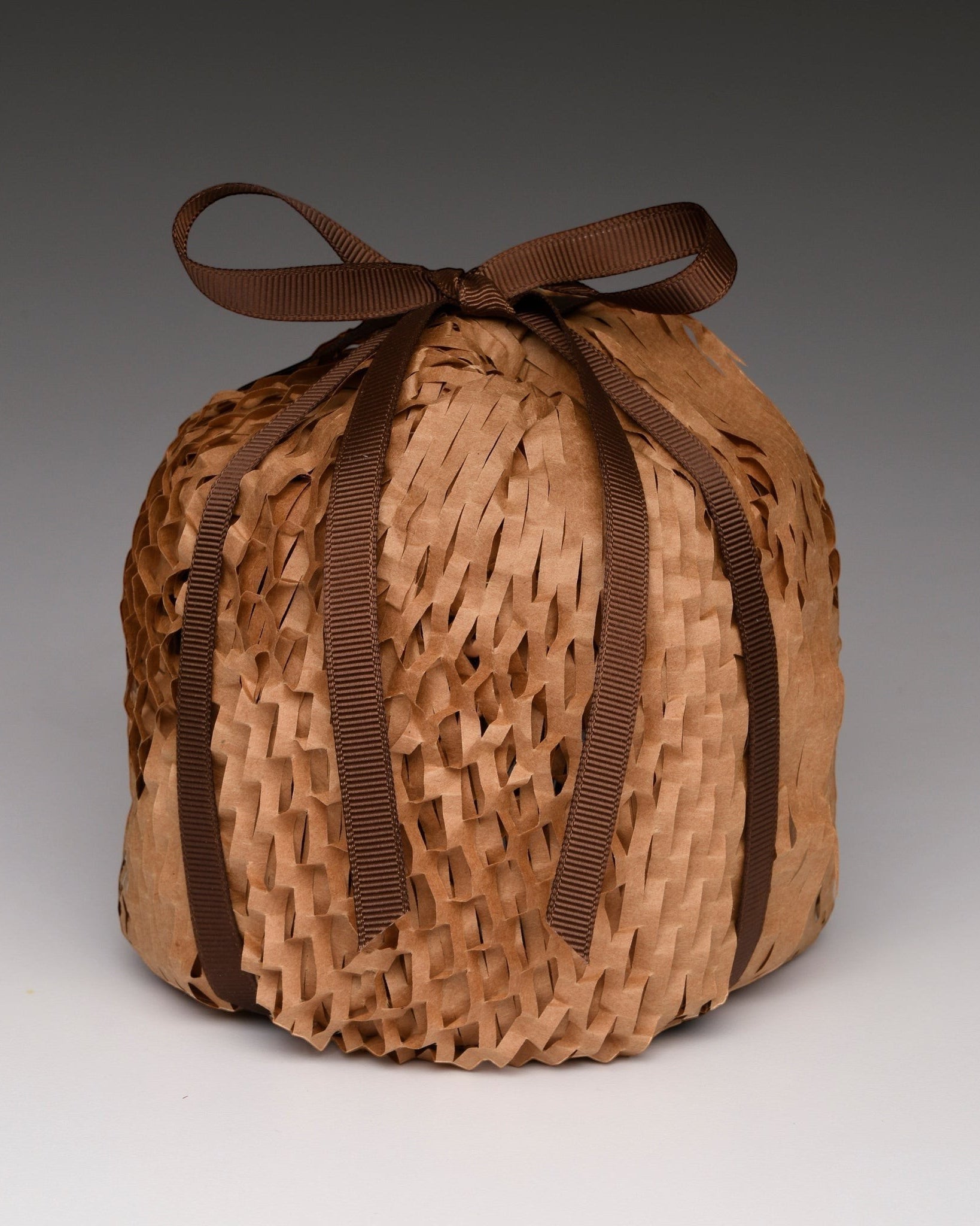 A round, hand-woven basket or pouch made of light brown natural fibers, tied at the top with a dark brown ribbon bow, set against a neutral gray gradient background.