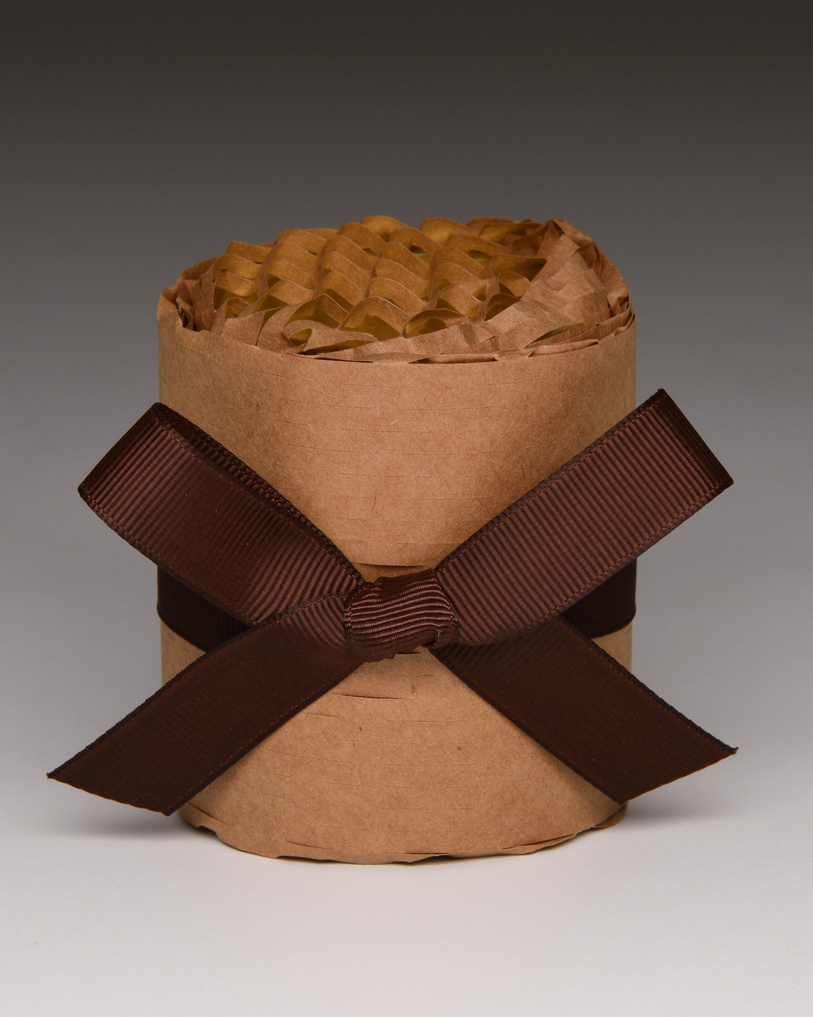 A brown kraft paper cylindrical container filled with crinkled brown tissue paper, tied with a wide dark brown ribbon bow in a studio setting.