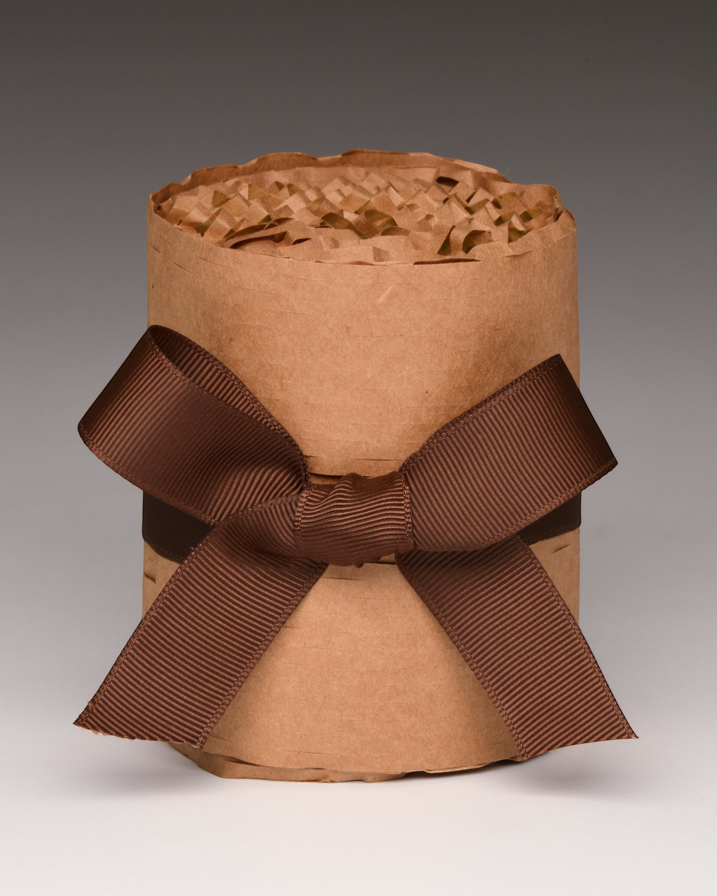 A brown kraft paper gift candle wrapped with a wide dark brown ribbon bow, filled with crinkle paper shred, photographed on a neutral gray gradient background.