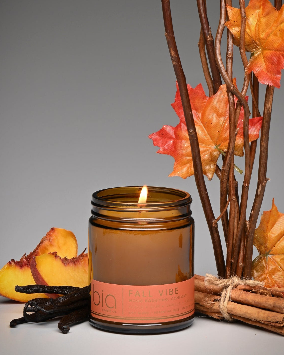 An amber glass jar candle with a lit flame and a peach-colored label reading 'KALY VIBE', styled with autumn leaves, cinnamon sticks, and vanilla pods on a gray background.