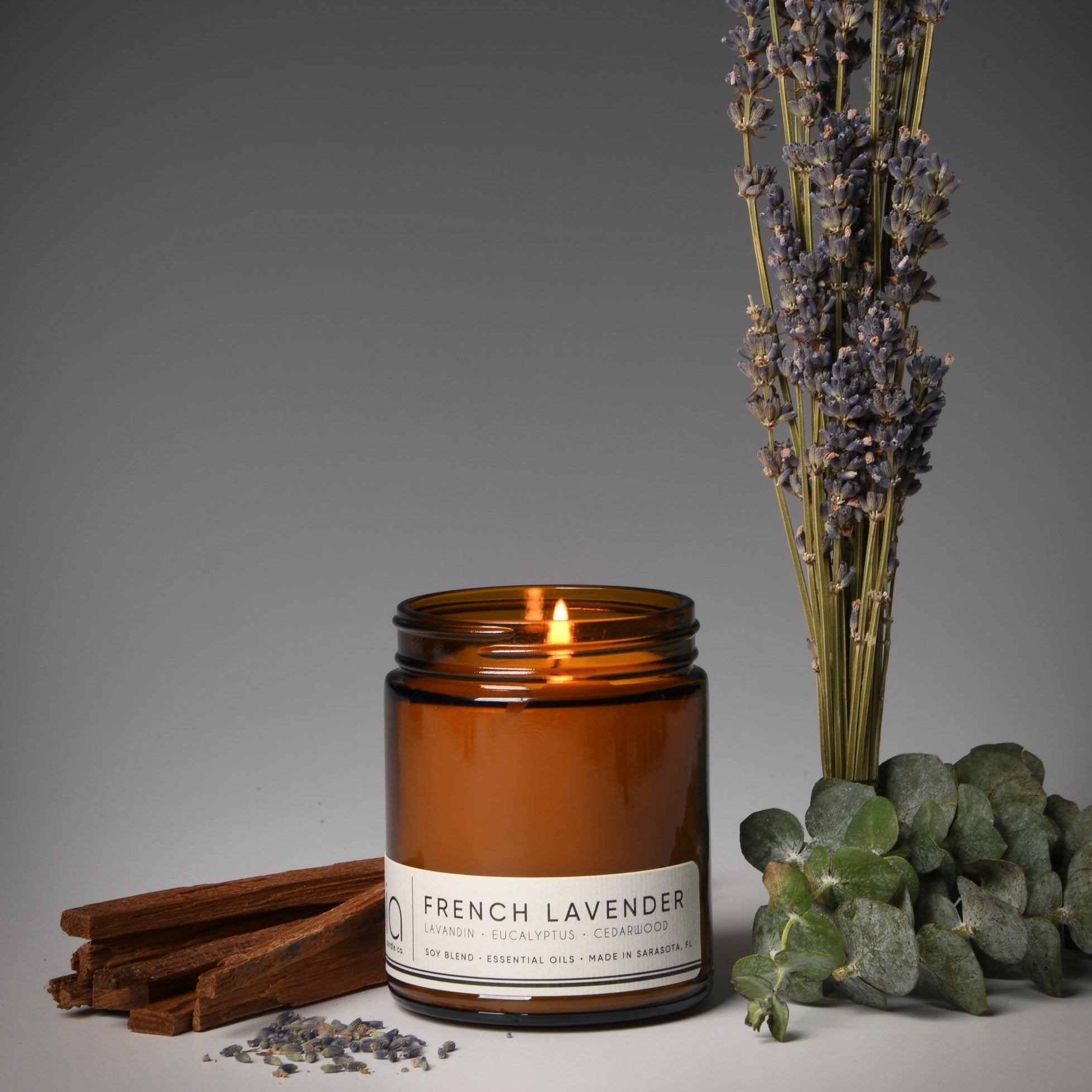 A lit French Lavender candle in a clear glass jar, with a background featuring a sprig of lavender and some cinnamon sticks.
