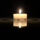 A single lit tea light candle in a clear plastic cup on a reflective surface, with a blurred background.