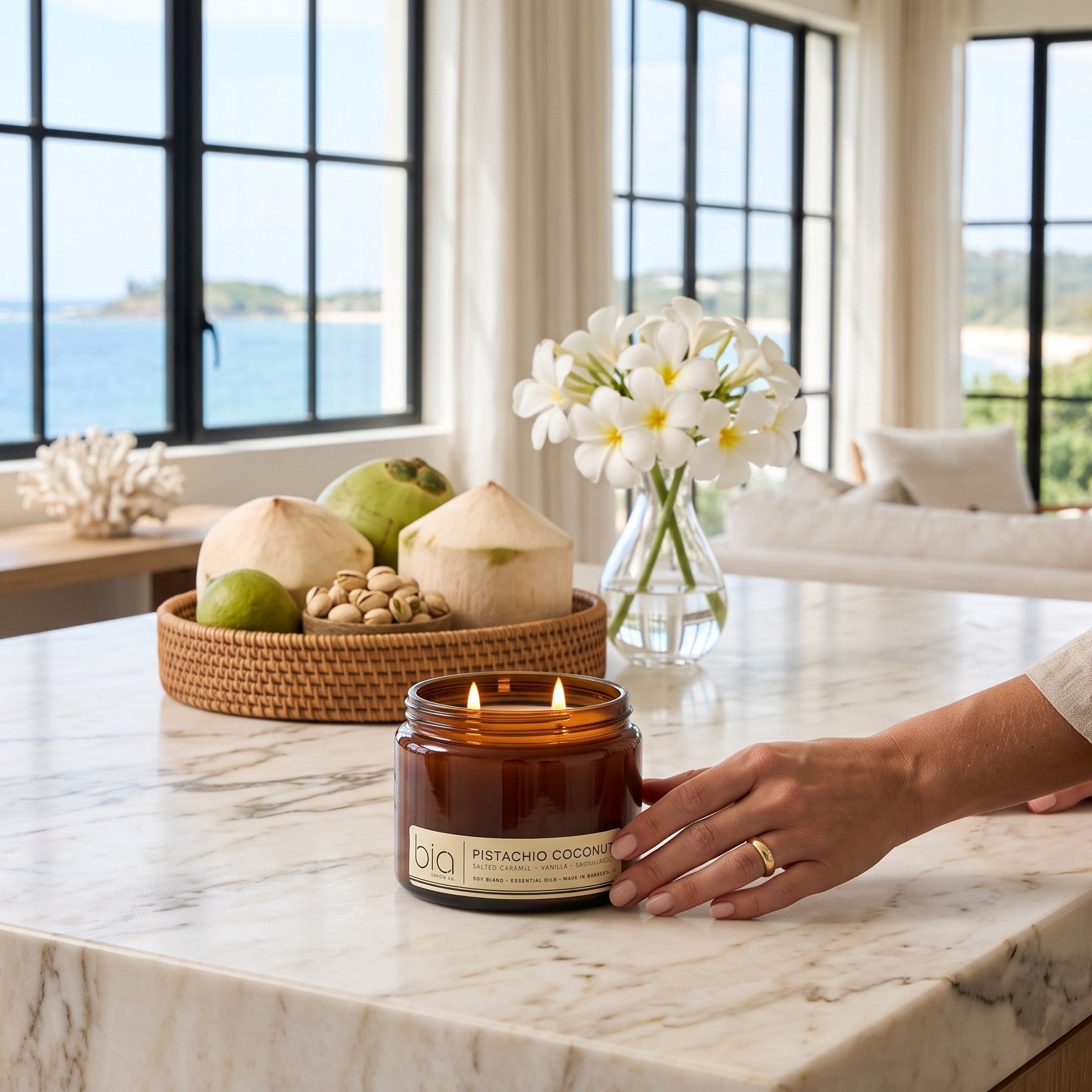 A lit amber glass jar candle labeled 'bia' sits on a white marble kitchen island, with a person's hand resting near it. The kitchen has large black-framed windows overlooking the ocean, with coconuts, flowers, and decorative items in the background.