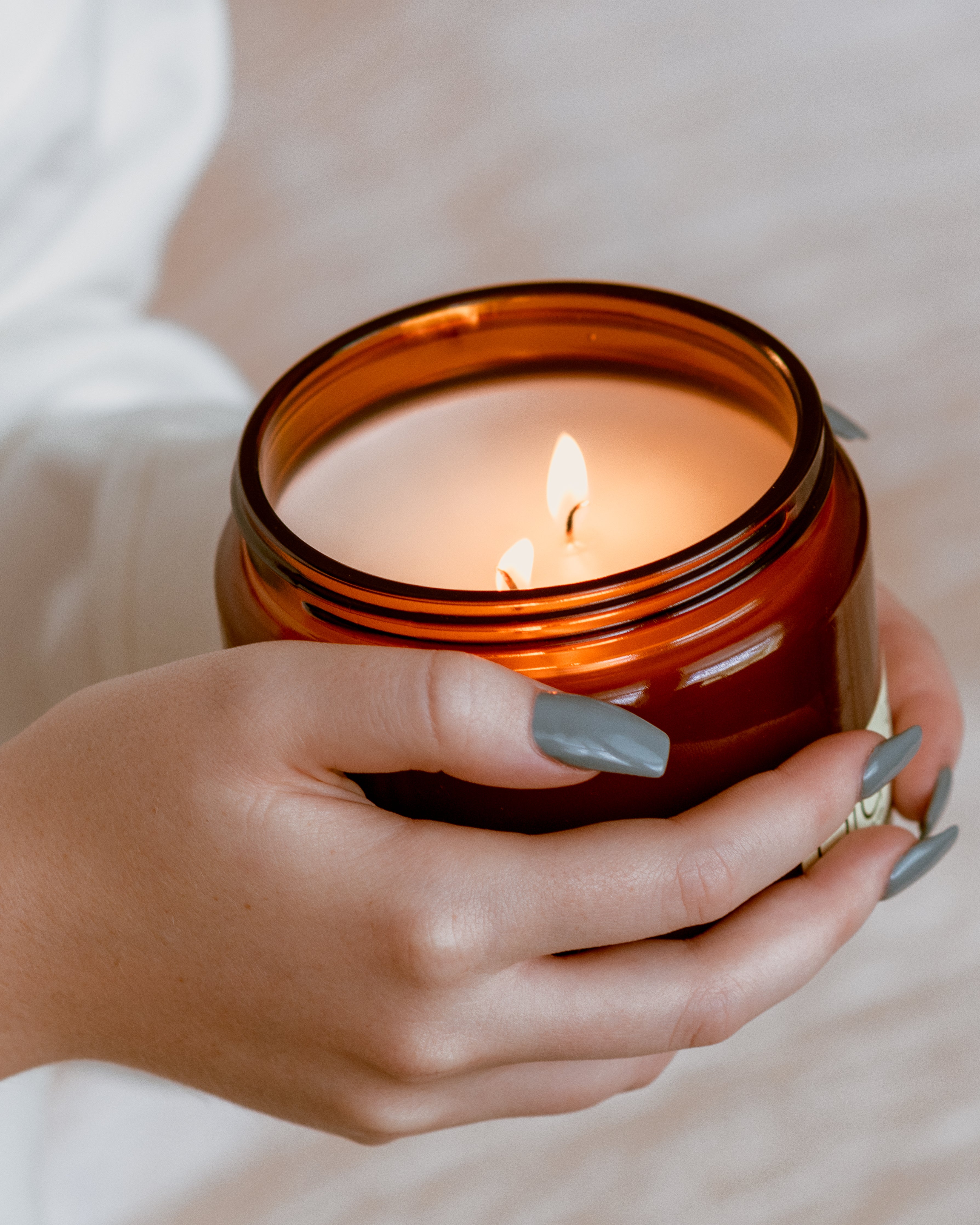 A lit amber glass jar candle held in two hands with gray manicured nails, set against a soft, light beige background.