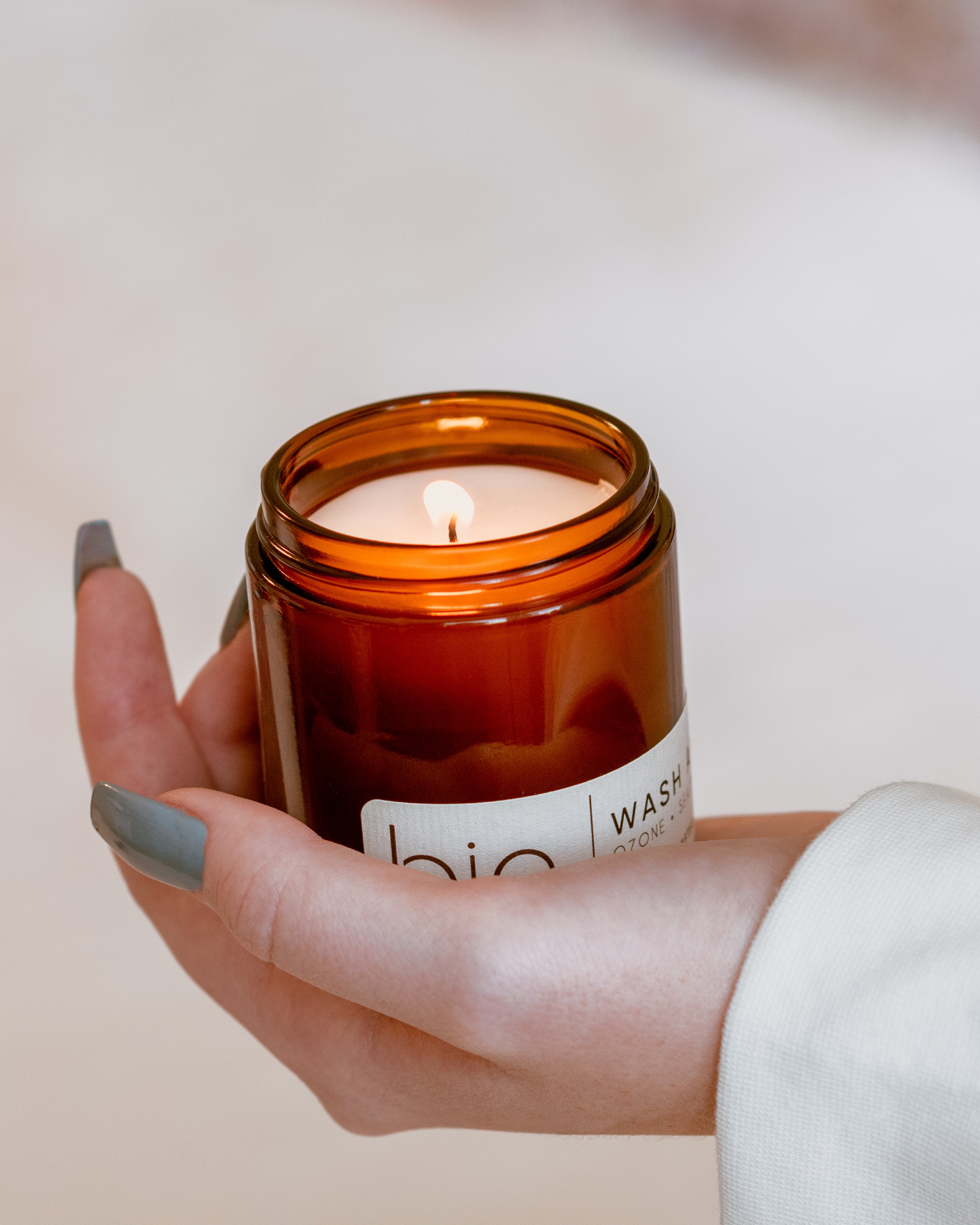 A lit amber glass jar candle held in a person's hand. The candle has a white label on brown jar