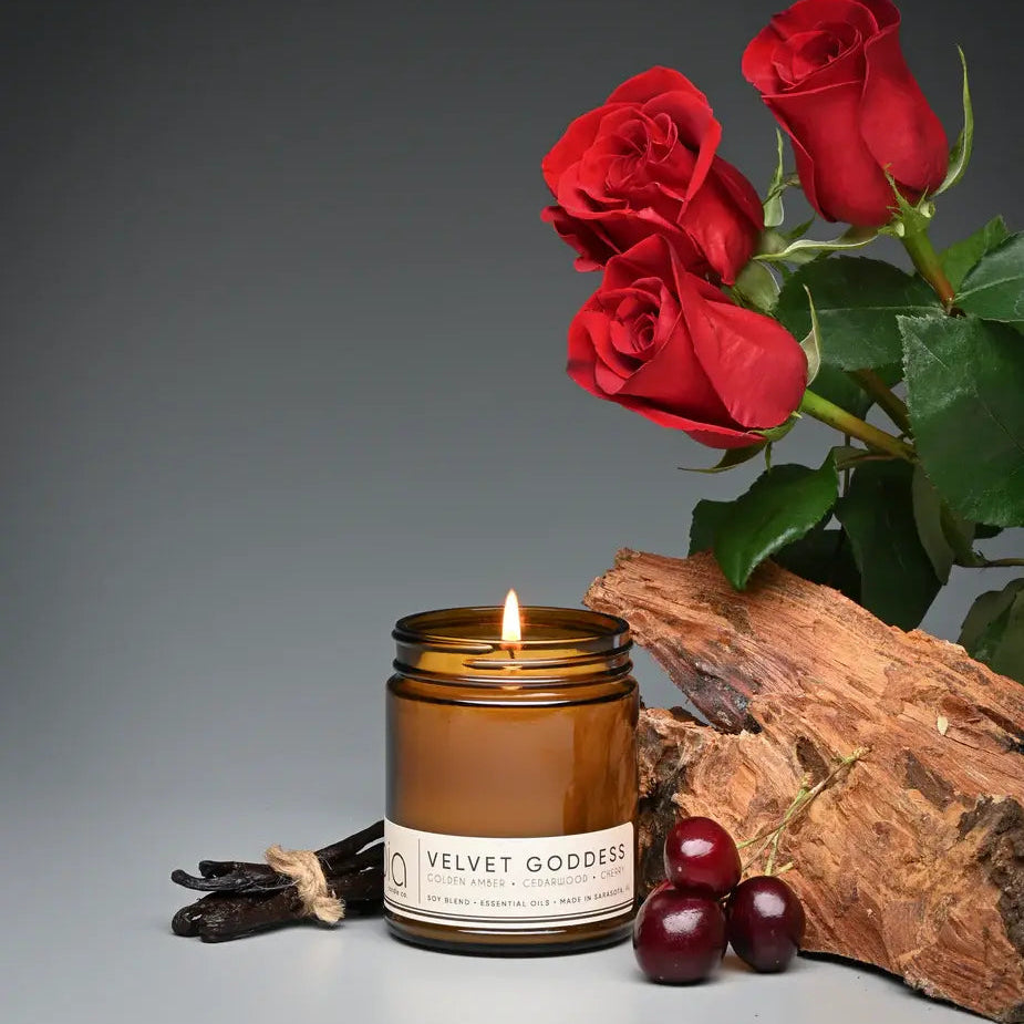 Candle labeled 'Velvet Goddess' with red roses, wood, and berries on a gray background