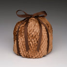 A round, hand-woven basket or pouch made of light brown natural fibers, tied at the top with a dark brown ribbon bow, set against a neutral gray gradient background.