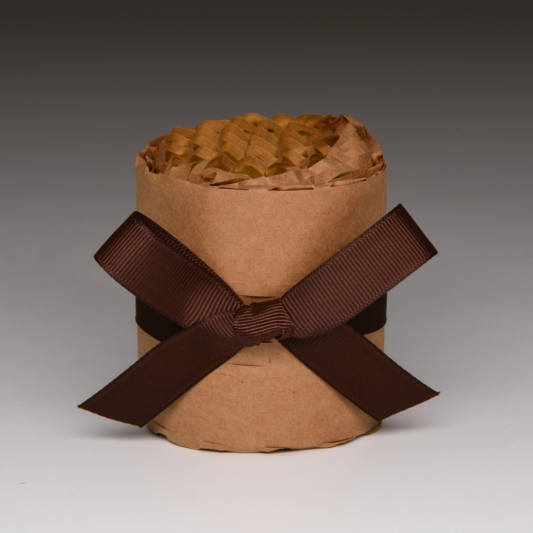 A brown kraft paper cylindrical container filled with crinkled brown tissue paper, tied with a wide dark brown ribbon bow in a studio setting.
