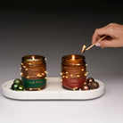 Two lit candles in decorative jars on a white tray with a hand lighting one of them using a match.