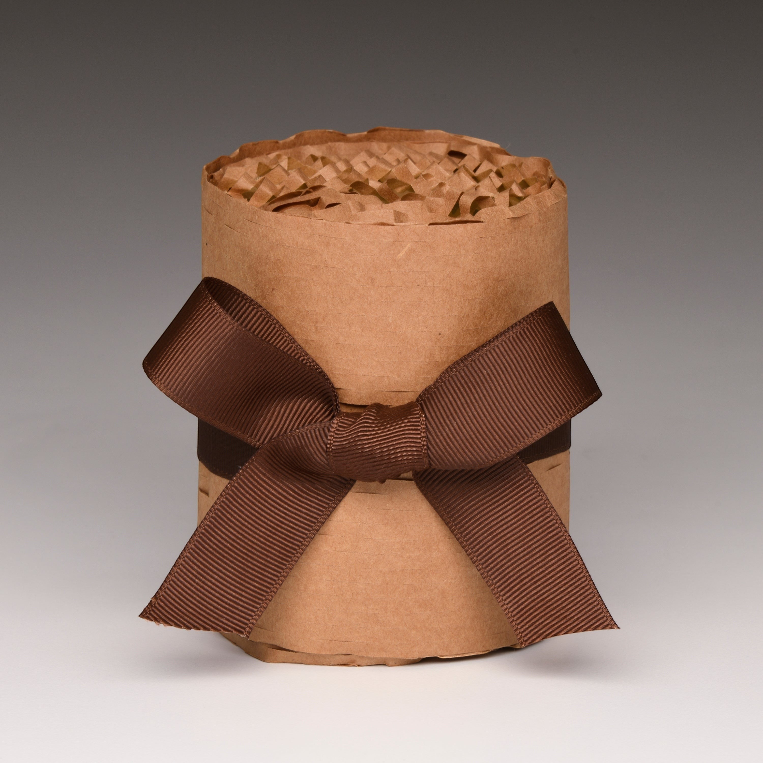 A brown kraft paper gift candle wrapped with a wide dark brown ribbon bow, filled with crinkle paper shred, photographed on a neutral gray gradient background.