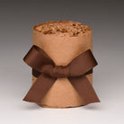 A brown kraft paper gift box wrapped with a wide dark brown ribbon bow, filled with crinkle paper shred, photographed on a neutral gray gradient background.