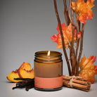 An amber glass jar candle with a lit flame and a peach-colored label reading 'KALY VIBE', styled with autumn leaves, cinnamon sticks, and vanilla pods on a gray background.