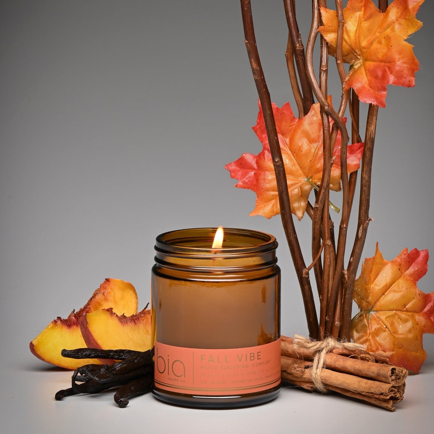 An amber glass jar candle with a lit flame and a peach-colored label reading 'KALY VIBE', styled with autumn leaves, cinnamon sticks, and vanilla pods on a gray background.