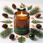 A lit amber classic glass candle jar labeled 'Fresh Woods' is arranged on a white surface with pine branches, pinecones, small amber beads, a green bow, and a green ornament.