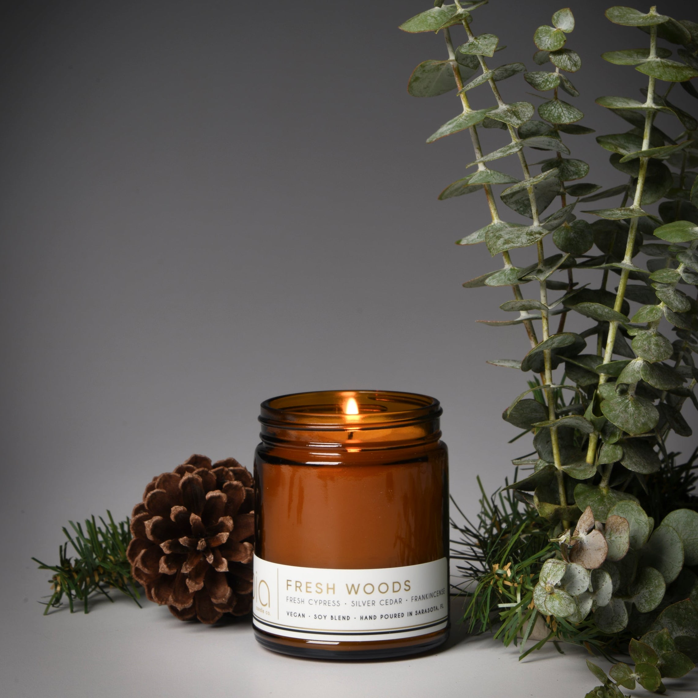 A lit candle in a clear glass jar with a brown label that reads 'FRESH WOODS'. It is surrounded by pine cones, pine needles, and green foliage against a gray background.