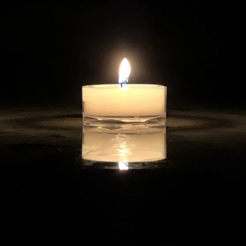 A single lit tea light candle in a clear plastic cup on a reflective surface, with a blurred background.