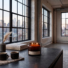 A lit amber glass jar candle sits on a dark countertop in a loft-style room with large black-framed windows overlooking a city skyline. A book, a small vase with dried grass, and two dark mugs are nearby.