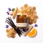 A lit amber glass jar candle labeled 'AMBER SANTAL' is arranged on a white background with orchid flowers, vanilla beans, cinnamon sticks, a slice of orange, and small purple blossoms.