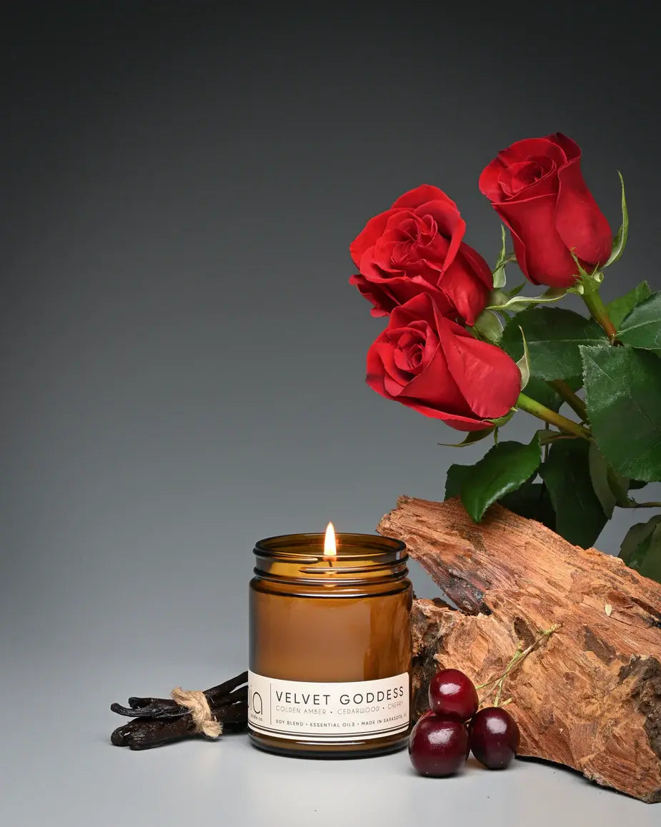 Candle labeled 'Velvet Goddess' with red roses, wood, and berries on a gray background